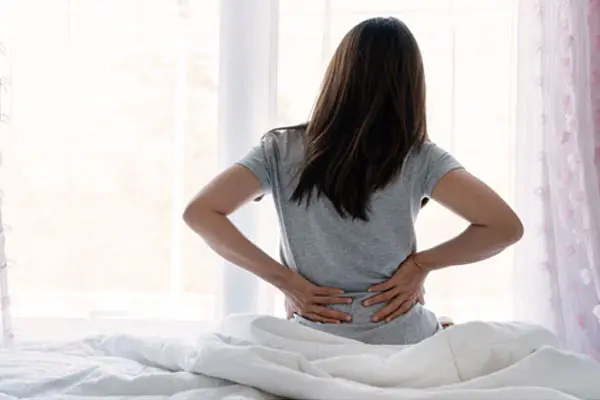 Mujer experimenta dolor de espalda mientras está sentada en la cama - SUNSGOODS - SUNSGOODS Mujer experimenta dolor de espalda mientras está sentada en la cama.